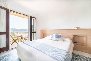 Une chambre d’hôtel avec un grand lit, une tête de lit en bois et une vue sur la mer, visible par une porte ouverte.