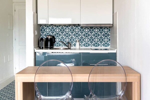 A compact kitchen with a coffee maker, a kettle, and a sink. There is a wooden table with two chairs in front.