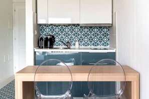 A compact kitchen with a coffee maker, a kettle, and a sink. There is a wooden table with two chairs in front.