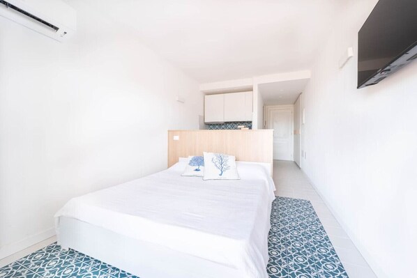 A bedroom with a bed, a nightstand, a wooden headboard, and a small kitchenette area.