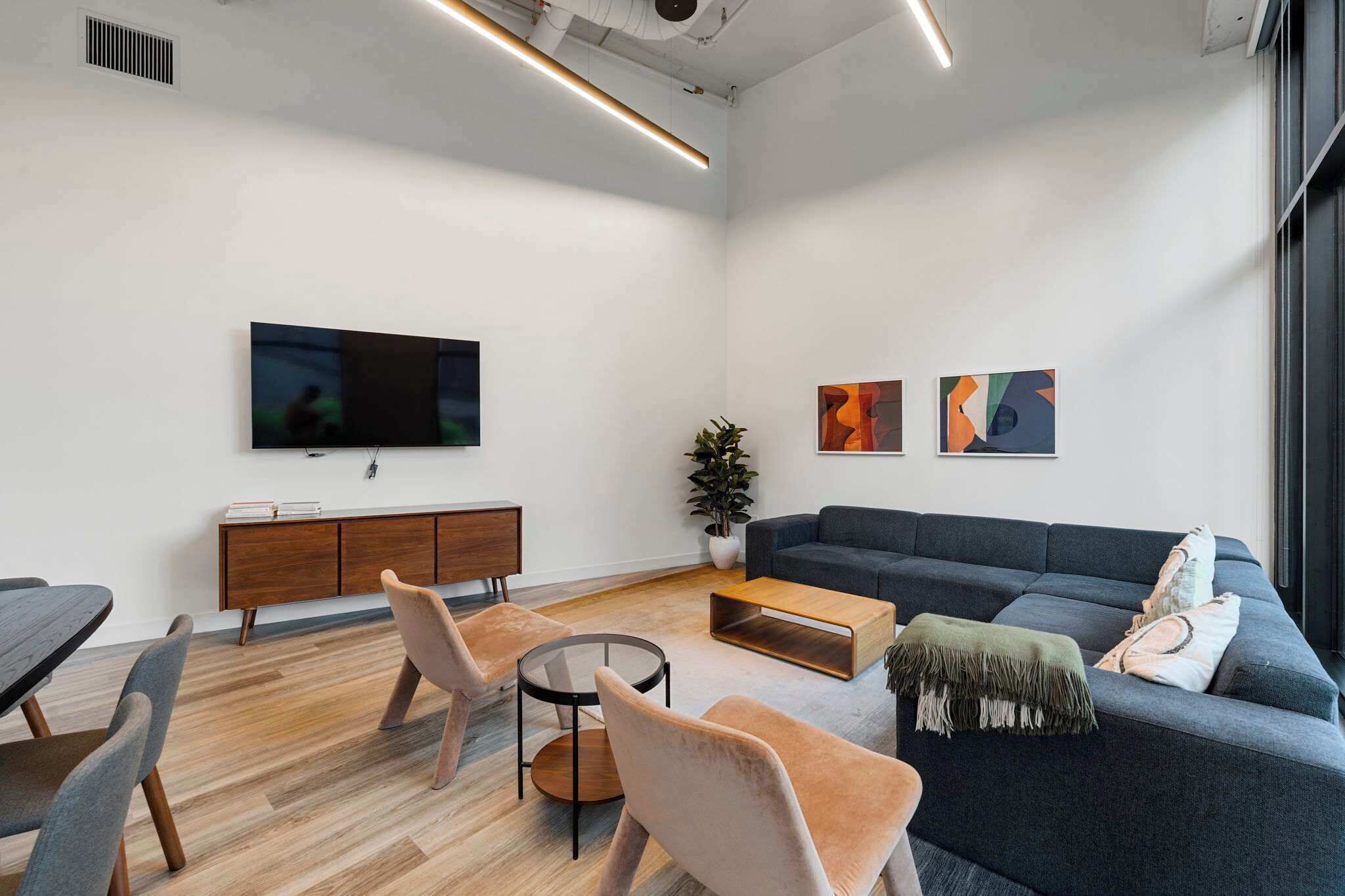 A modern living room with a flat-screen TV, a dark sofa, a coffee table, and a dining area with chairs and a wooden cabinet.