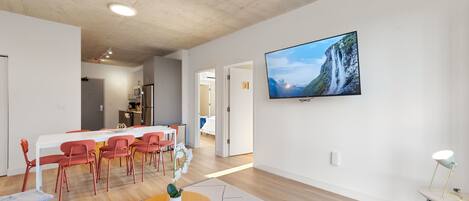 A modern living room with a dining area, a flat-screen TV mounted on the wall, and a kitchen in the background.