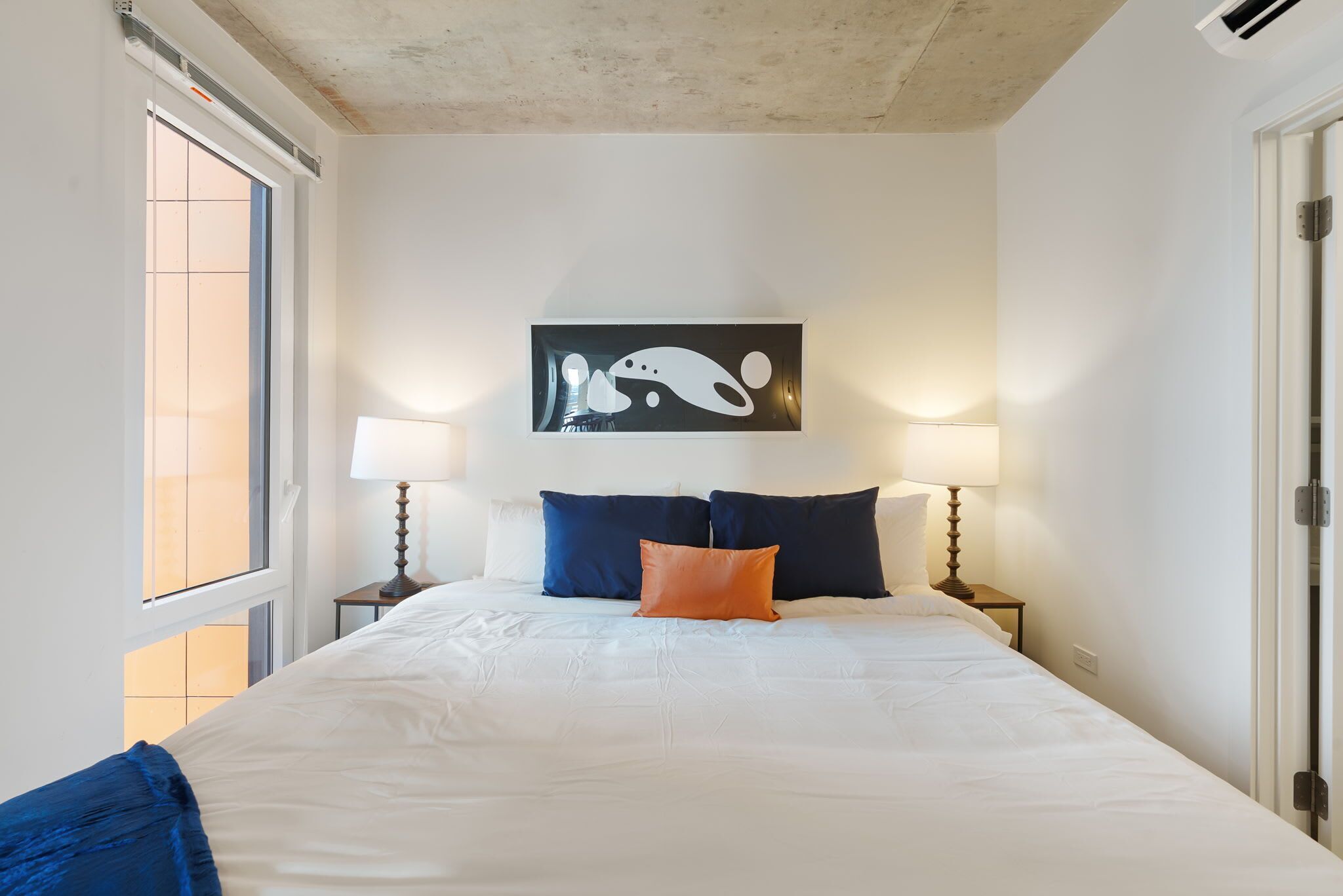 A hotel room with a large bed, two bedside lamps, a window, and a wall-mounted artwork.