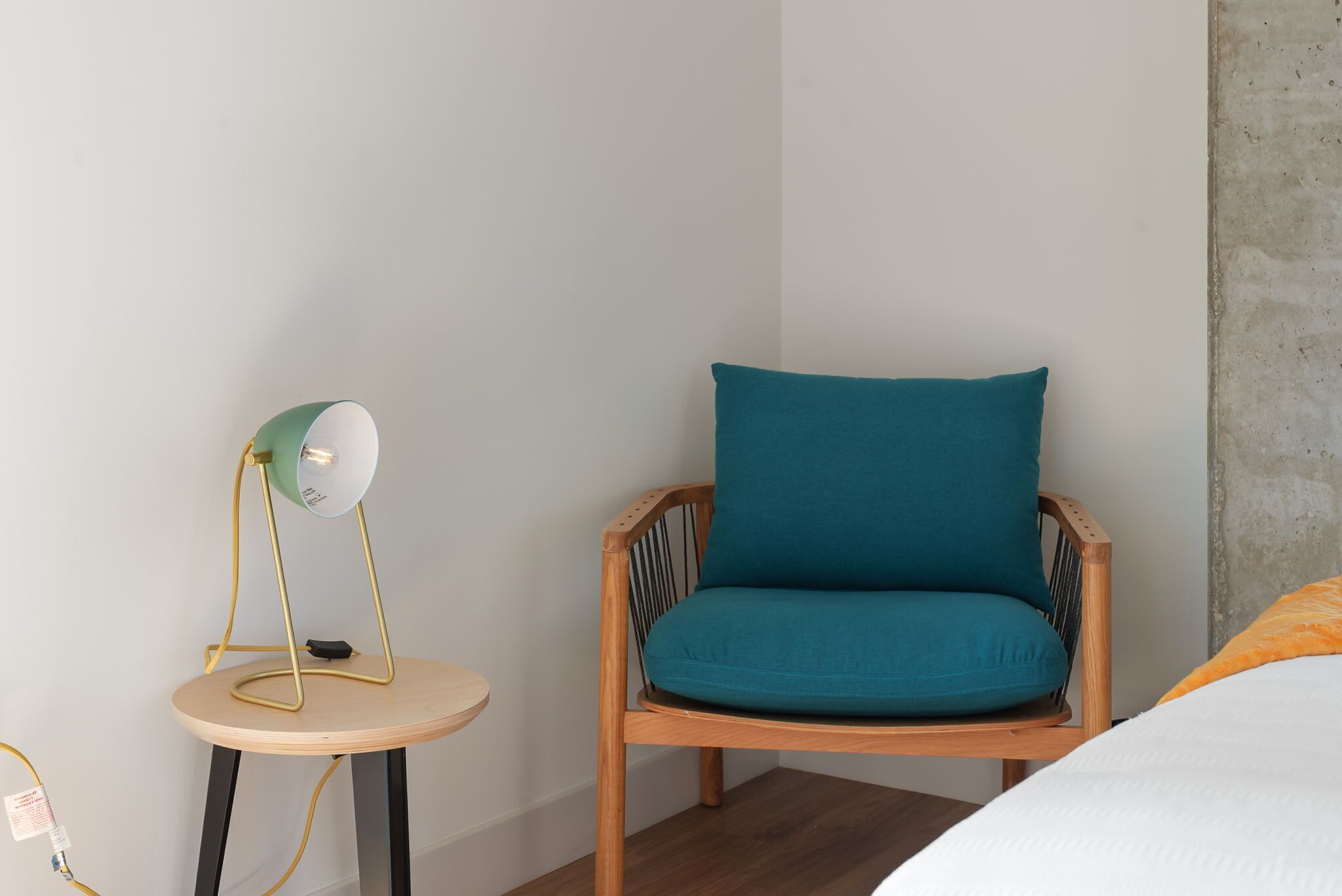 A room with a wooden chair with teal cushions, a small round wooden table with a green lamp, and a white bed with an orange blanket.