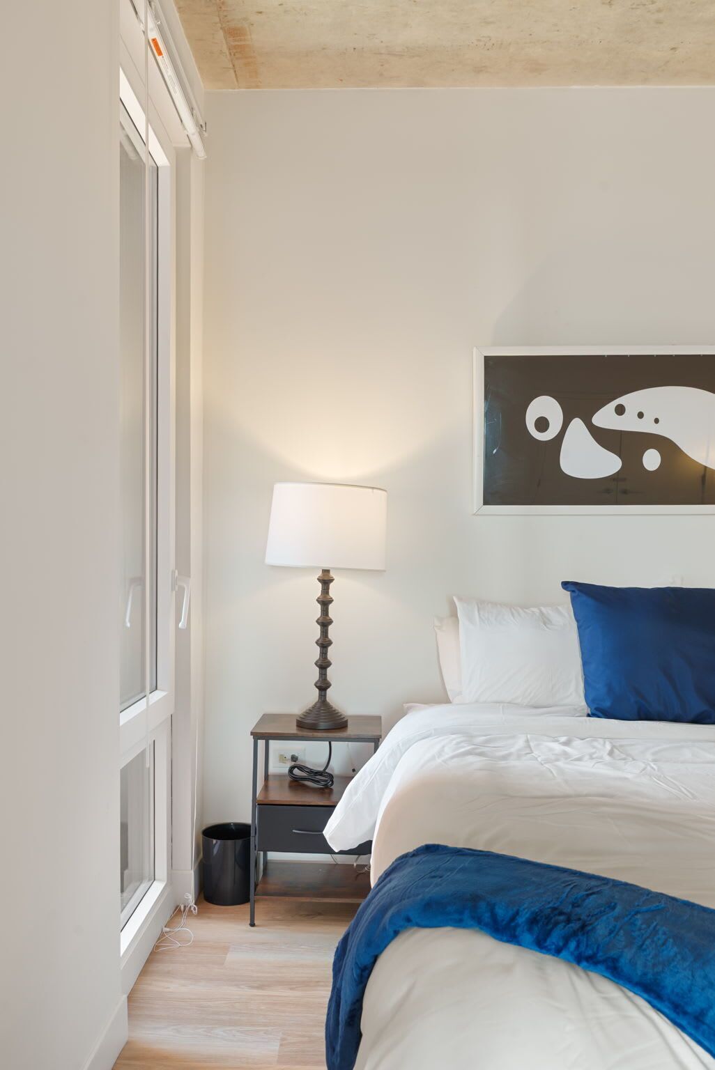 A bedroom with a bed, a nightstand with a lamp, a trash bin, and a wall art piece.