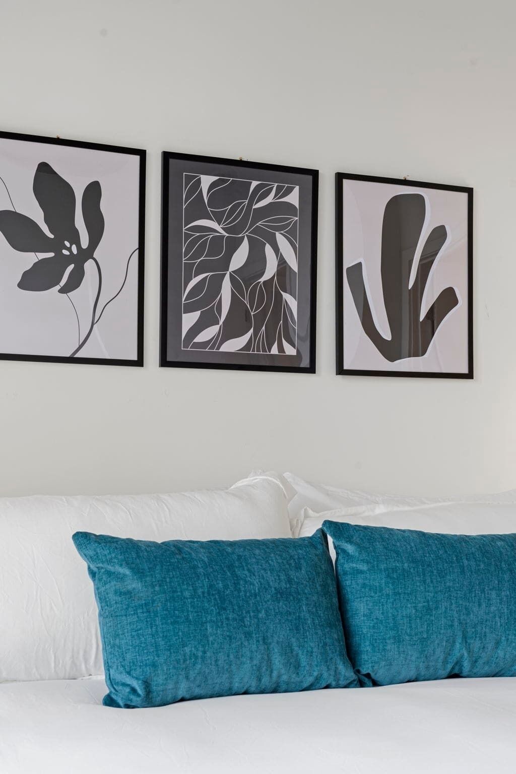 A bedroom with a white bed, teal pillows, and three framed abstract art pieces on the wall.