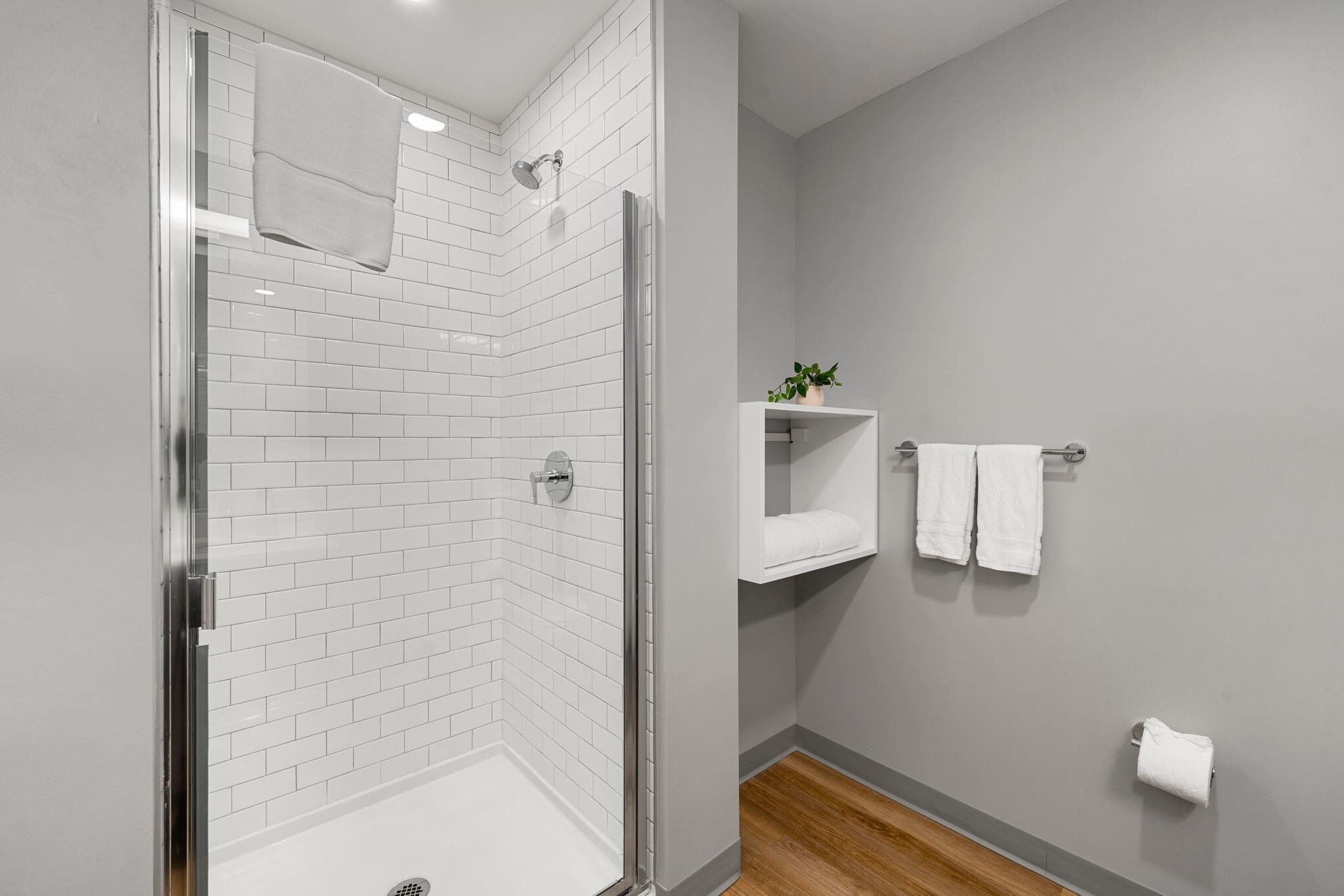 A modern bathroom with a glass-enclosed shower, white tiled walls, a wooden floor, and a small shelf with a plant.