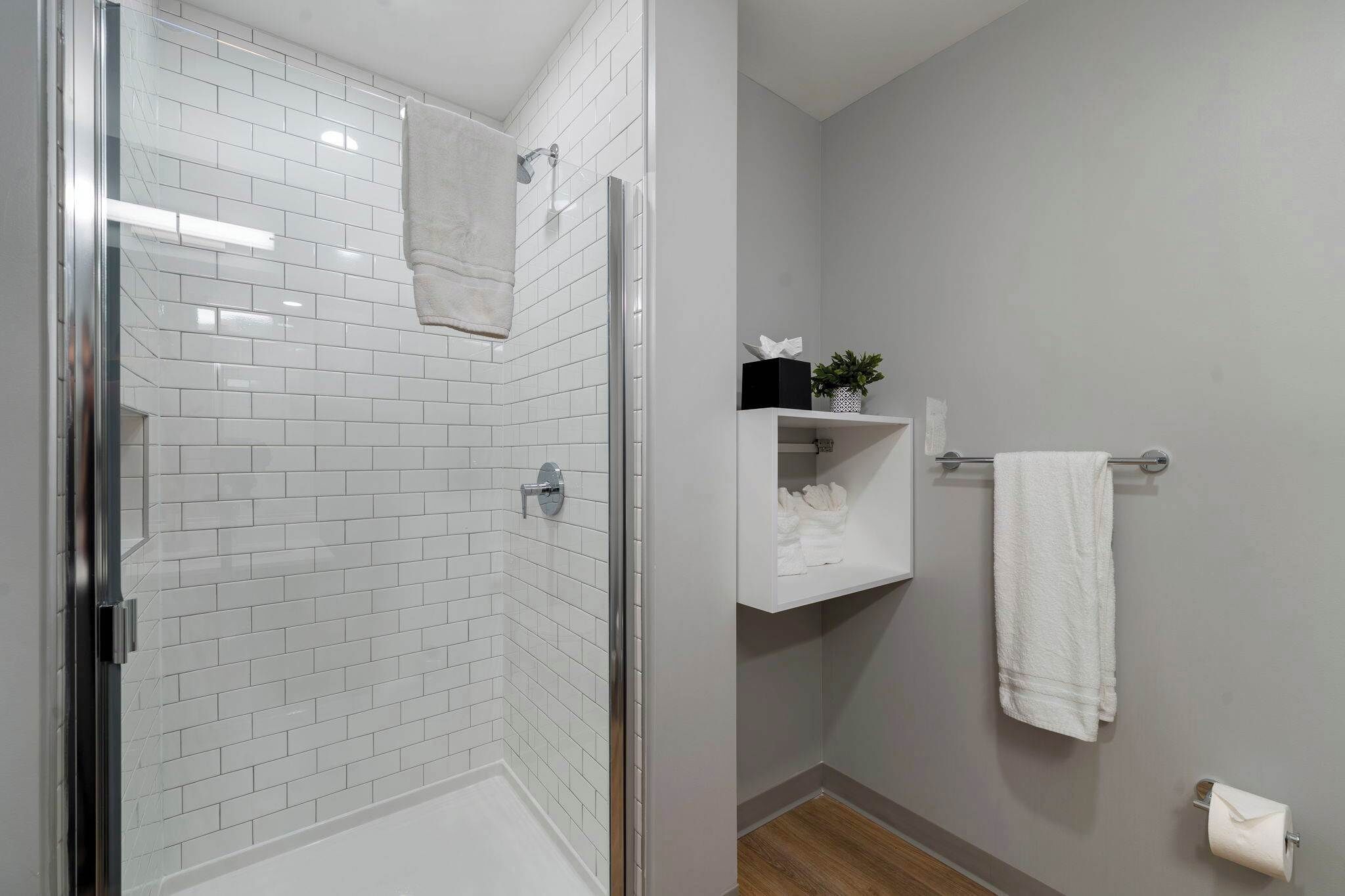 A bathroom with white tiled walls, a glass shower enclosure, a white towel hanging on a rack, and a small shelf with toiletries.