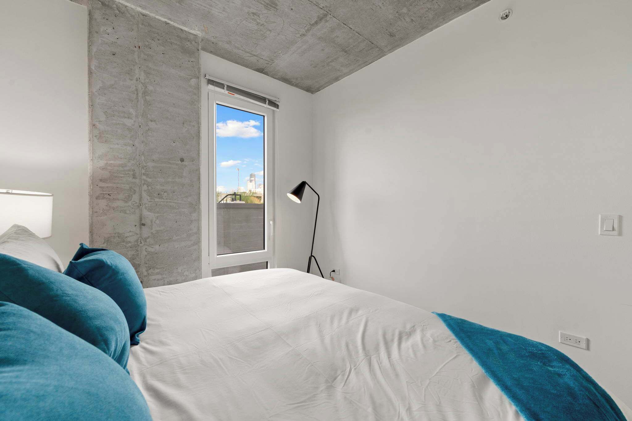 Un dormitorio con cama, ventana, lámpara y pared de concreto.