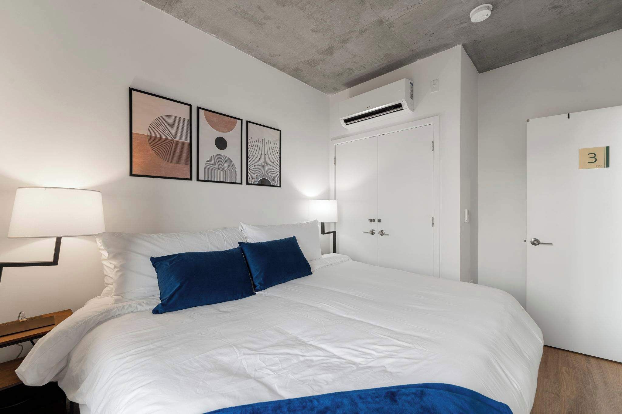 Habitación de hotel con una cama grande, dos lámparas de noche, un aire acondicionado instalado en la pared y una puerta con el número indicado.