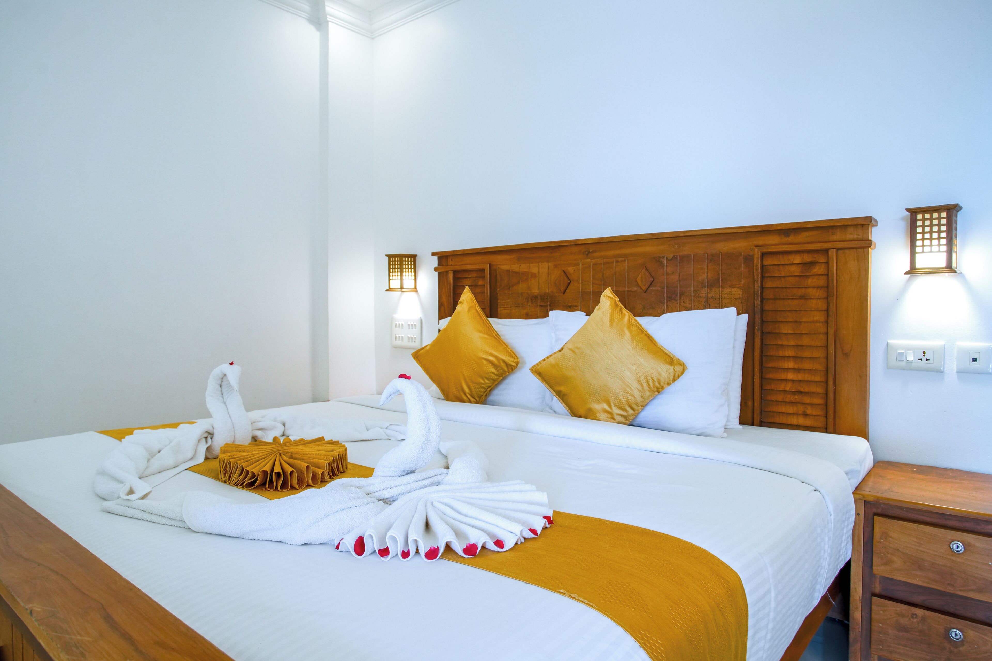 A hotel room with a neatly made bed featuring a swan towel arrangement, a wooden headboard, and two wall-mounted lights.