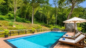 Una piscina rectangular con borde de azulejos, rodeada por una terraza de madera. Hay sillones de descanso con sombrillas y una pequeña mesa con una planta.