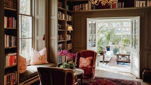 A library with bookshelves, a reading chair, a red armchair, and a view through an open door.