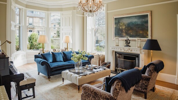 A spacious living room with a grand chandelier, a fireplace, and a large window offering a view of the city.