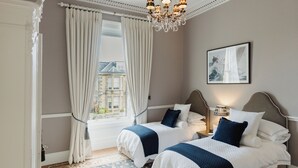 A hotel room with two beds, a large window with curtains, a chandelier, and a framed picture on the wall.