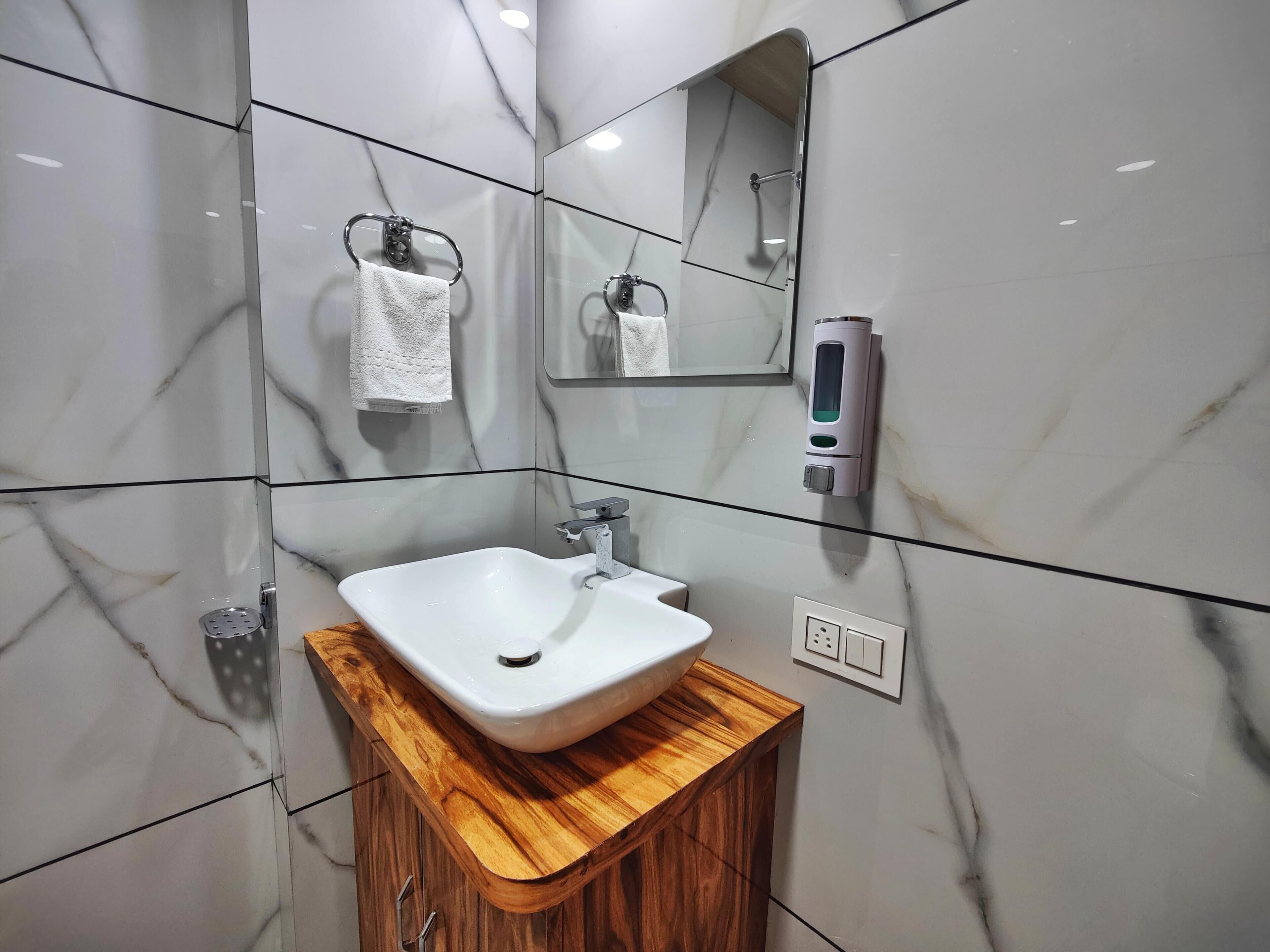 A bathroom with a white sink, wooden vanity, marble tiles, a towel rack, a mirror, and a soap dispenser.