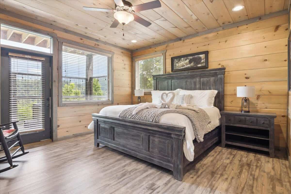 Une chambre avec un plafond en bois, un lit doté d’une tête de lit sombre, deux tables de chevet avec des lampes et un fauteuil à bascule.