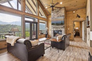A cozy cabin interior with a fireplace, leather sofas, and large windows offering a scenic view. - Spacious 3BR Gatlinburg Cabin w/ Hot Tub, Fire Pit, & Game Room, Great Location (Gatlinburg)