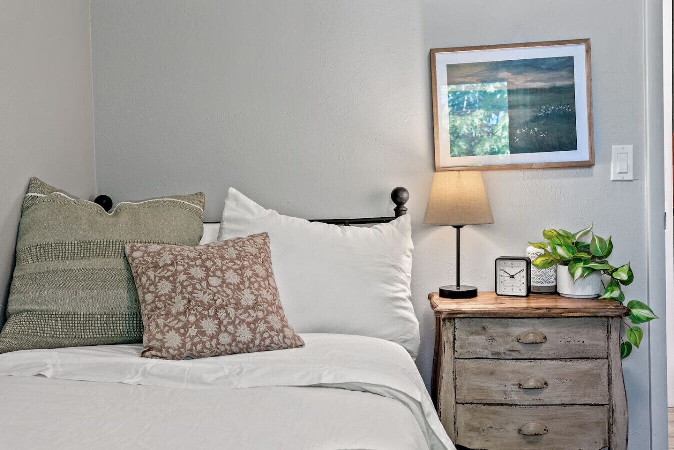Une chambre à coucher avec un lit, des oreillers, une table de chevet avec une lampe, une horloge et un tableau encadré au mur.