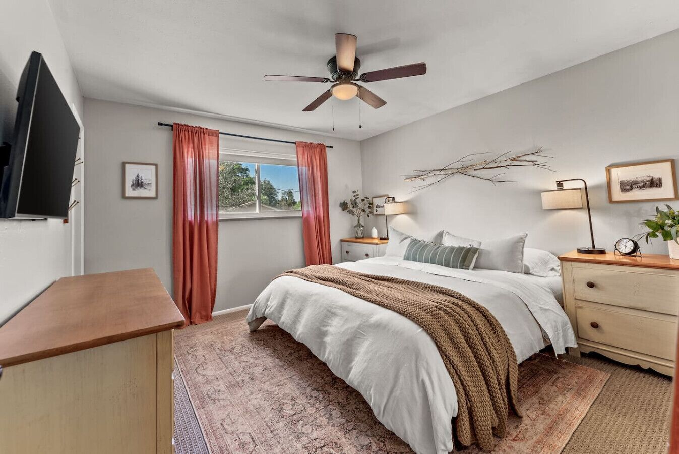 Quarto com cama, mesa de cabeceira, escrivaninha, ventoinha no teto, janela com cortinas e televisão fixada na parede.