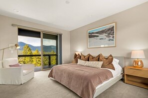 A bedroom with a large bed, a chair, a nightstand, a lamp, and a window with a mountain view. - Summit Solace | LUXE 360° Views • Hot Tub • Games (Bailey)
