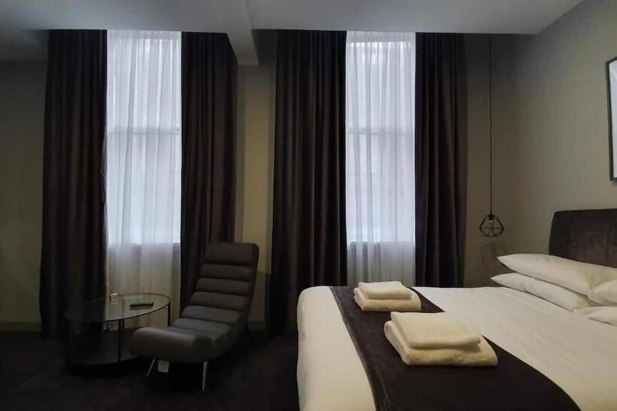 Une chambre d’hôtel avec un lit, une chaise, une petite table et de grandes fenêtres avec des rideaux.