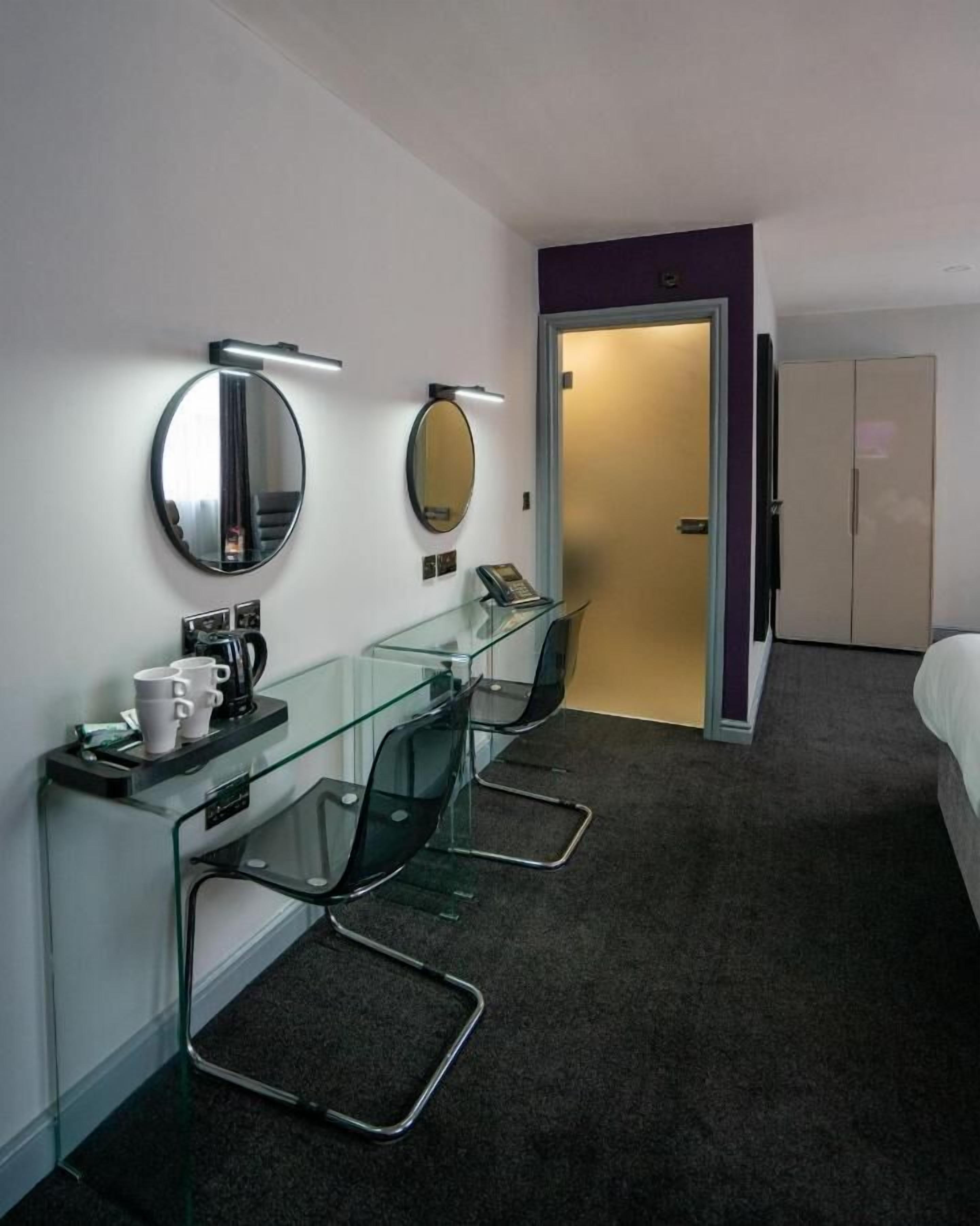 Ein Hotelzimmer mit Glastisch, zwei Stühlen, einem Bett, einem Kleiderschrank und einer Tür zu einem weiteren Raum.