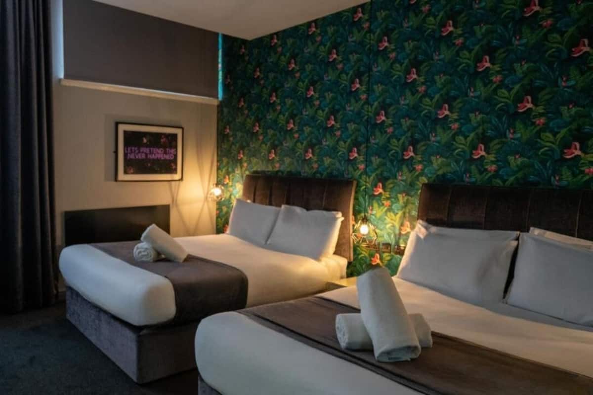Une chambre d’hôtel avec deux lits, une grande fenêtre avec des rideaux, un mur recouvert d’un papier peint aux motifs tropicaux et une citation encadrée accrochée au mur.