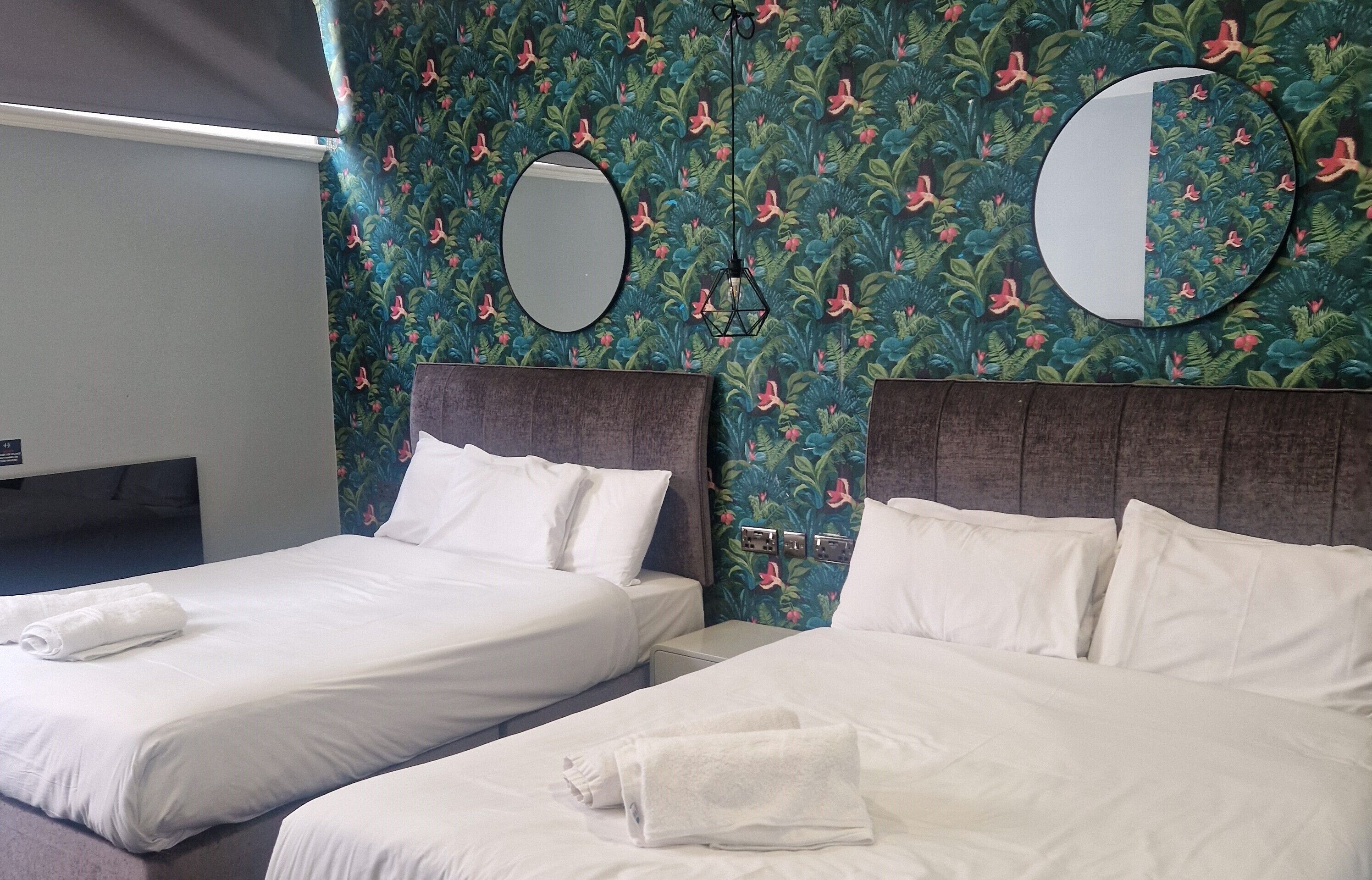 Two single beds with white linens and towels, a round mirror above each bed, and a floral-patterned wall.