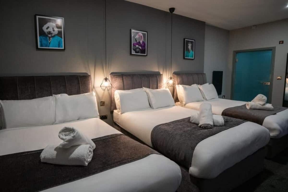 Une chambre d’hôtel avec trois lits, chacun doté de draps blancs et d’une tête de lit sombre. Des appliques murales et des tableaux encadrés sont fixés au mur au-dessus des lits.