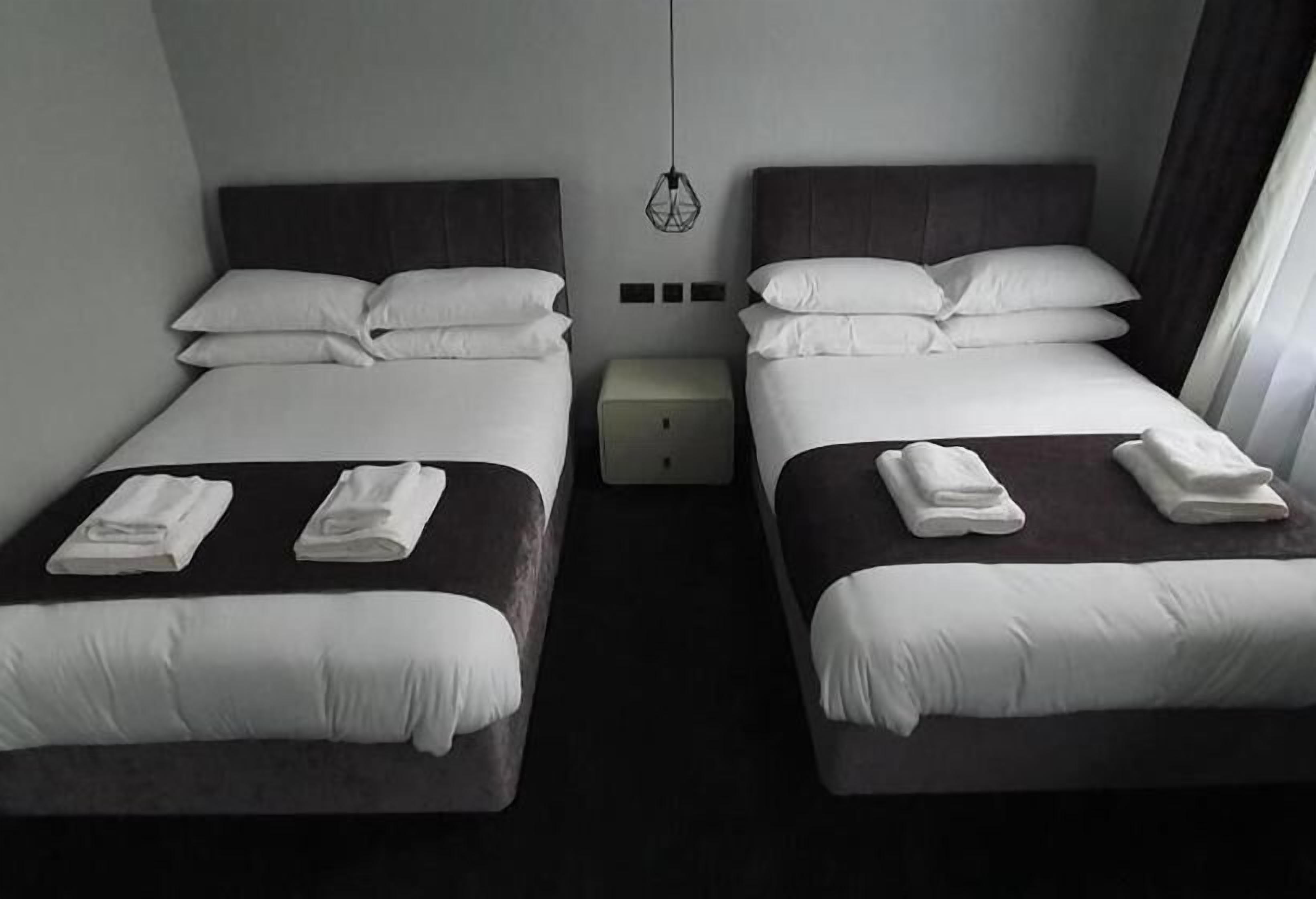 A hotel room with two beds, each with white bedding and pillows, a small bedside table, and a hanging light fixture.