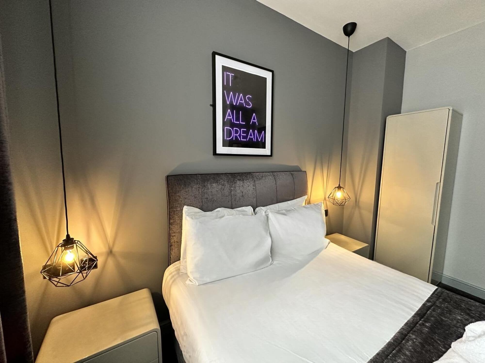 A modern hotel room with a bed, two bedside lamps, a wardrobe, and a neon sign on the wall.