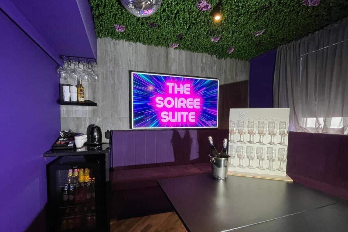 Une pièce avec un panneau néon indiquant « THE SOIREE SUITE », un coin bar avec des verres et des bouteilles, et un mur vert orné d’une plante.