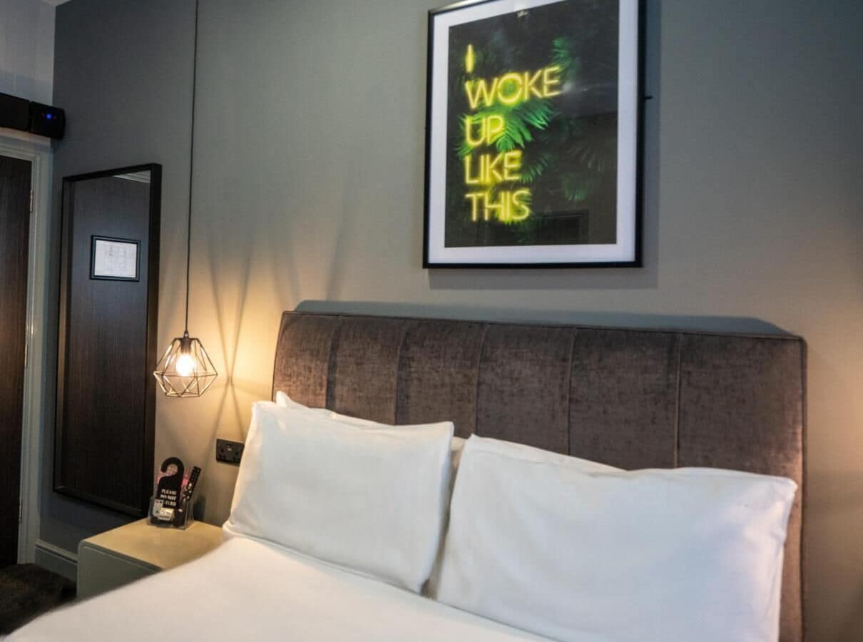A hotel room with a bed, a nightstand with a lamp, a framed motivational quote on the wall, and a door.