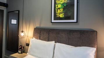 A hotel room with a bed, a nightstand with a lamp, a framed motivational quote on the wall, and a door.