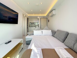 Camera da letto moderna con un letto, comodini, televisore e angolo cottura visibile attraverso una porta aperta.