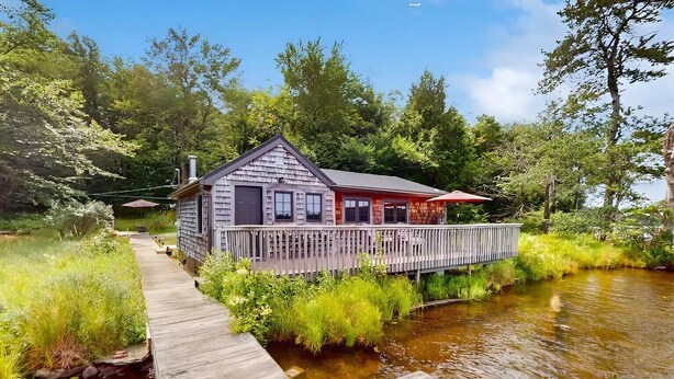 Une cabane dotée d’une terrasse et d’un petit quai s’avançant sur un ruisseau, le tout entouré d’une végétation luxuriante.