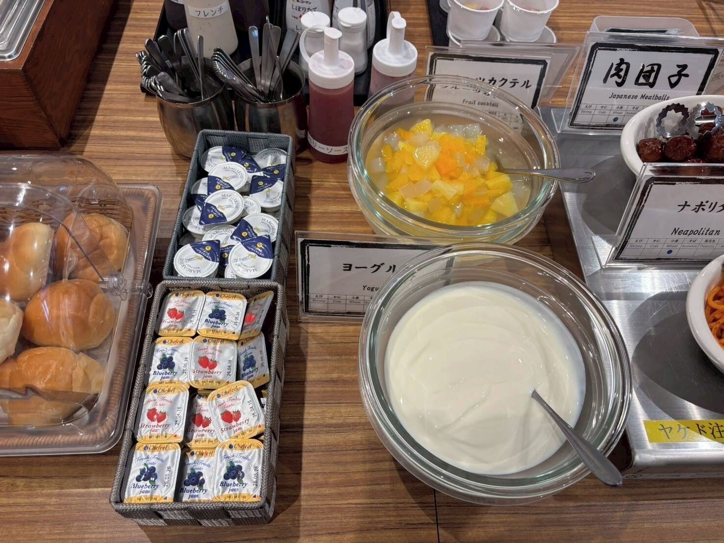 Verschiedene Speisen und Würzmittel auf einem Tisch, darunter abgepackte Marmeladen, eine Fruchtbowl und ein Joghurtbecher.