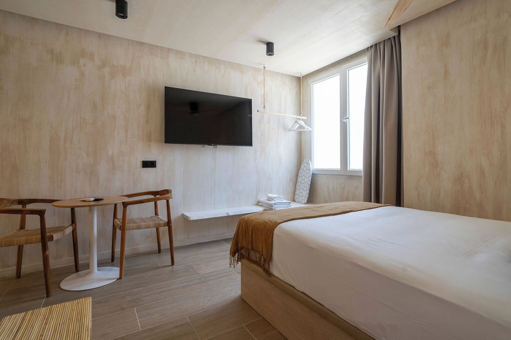 Une chambre d’hôtel avec un lit, un téléviseur fixé au mur, une petite table avec des chaises et une fenêtre avec des rideaux.
