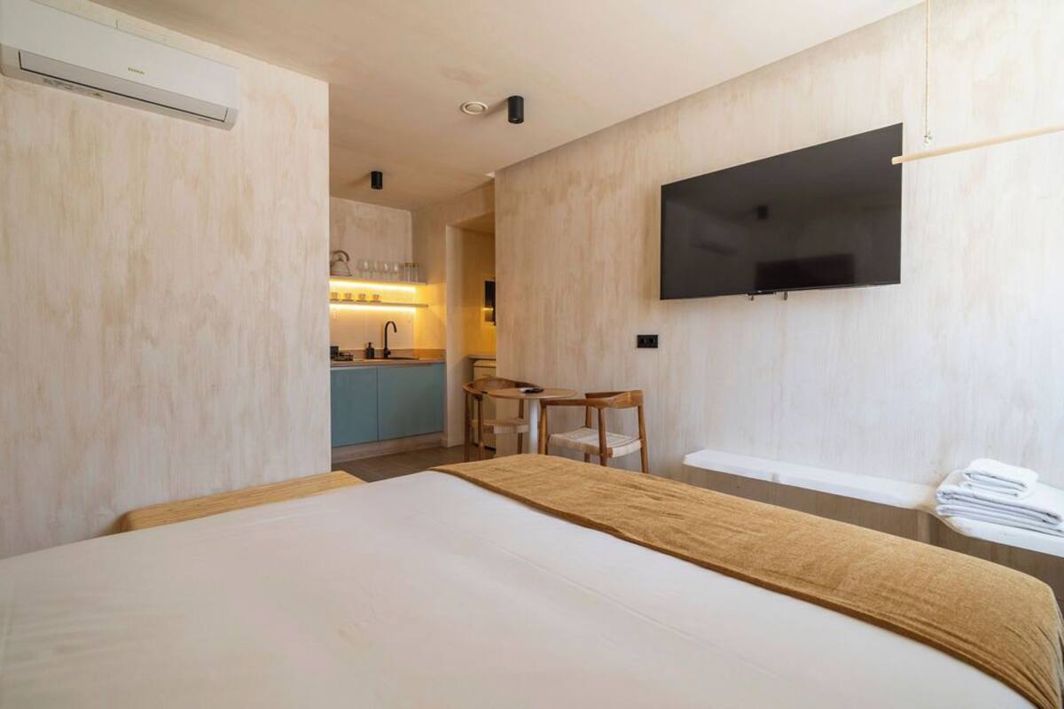Een hotelkamer met een bed, een aan de muur bevestigde televisie, een kleine tafel met stoelen en een kitchenette op de achtergrond.