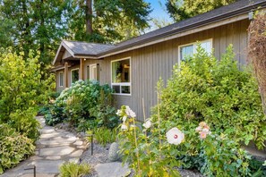 Une maison en bois avec un chemin de pierre menant à l’entrée, entourée d’une végétation luxuriante et de plantes en fleurs.