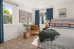 Une chambre à coucher avec un lit, une chaise, une petite table sur laquelle se trouve une plante, une lampe et deux tableaux encadrés accrochés au mur.