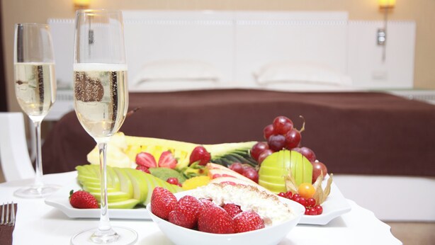 Una mesa de comedor con una bandeja de frutas, dos copas de champán y una cama al fondo.