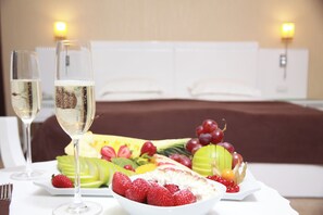 A dining table with a fruit platter, two glasses of champagne, and a bed in the background. - Amber Hotel (Baku)