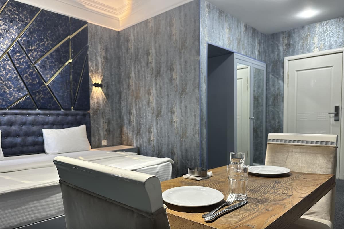 A modern hotel room with a large bed, a wooden dining table set with plates and cutlery, and a built-in headboard with a geometric design.