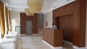 Un vestíbulo de hotel con un mostrador de recepción, una zona de espera con sofás blancos y una escalera.
