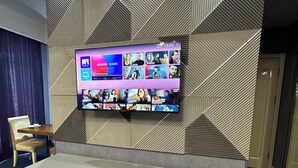 Habitación de hotel con una pared de diseño geométrico, un televisor de pantalla plana, un escritorio con silla y un pequeño cesto de basura.