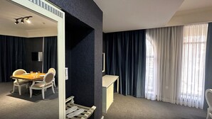 Una habitación de hotel moderna con zona de comedor, un escritorio pequeño y un ventanal con cortinas.