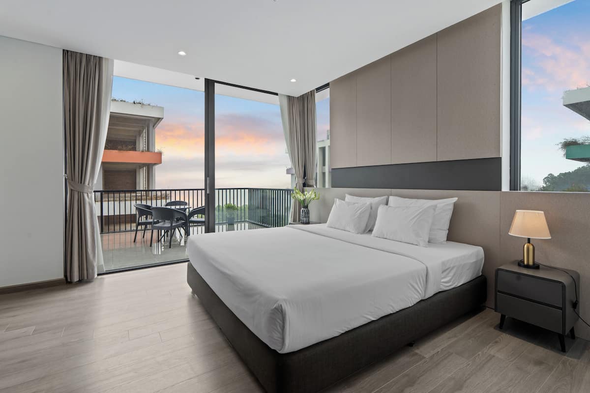 A modern hotel room with a large bed, a nightstand, a desk with a chair, and a balcony with a view of the sunset.