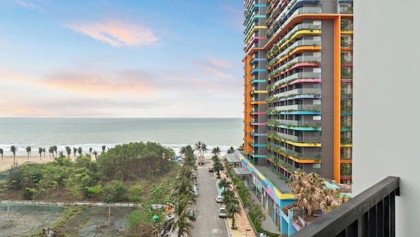 A high-rise hotel with balconies, a beach view, and a road with palm trees.